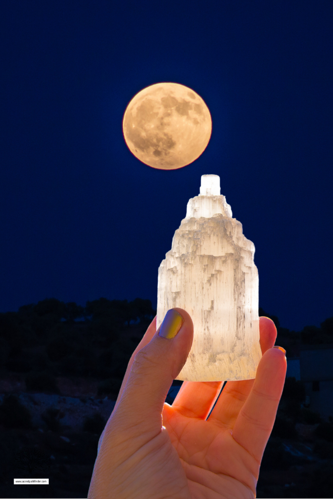 Photo of a hand holding up a selenite crystal in front of a full moon to charge it.