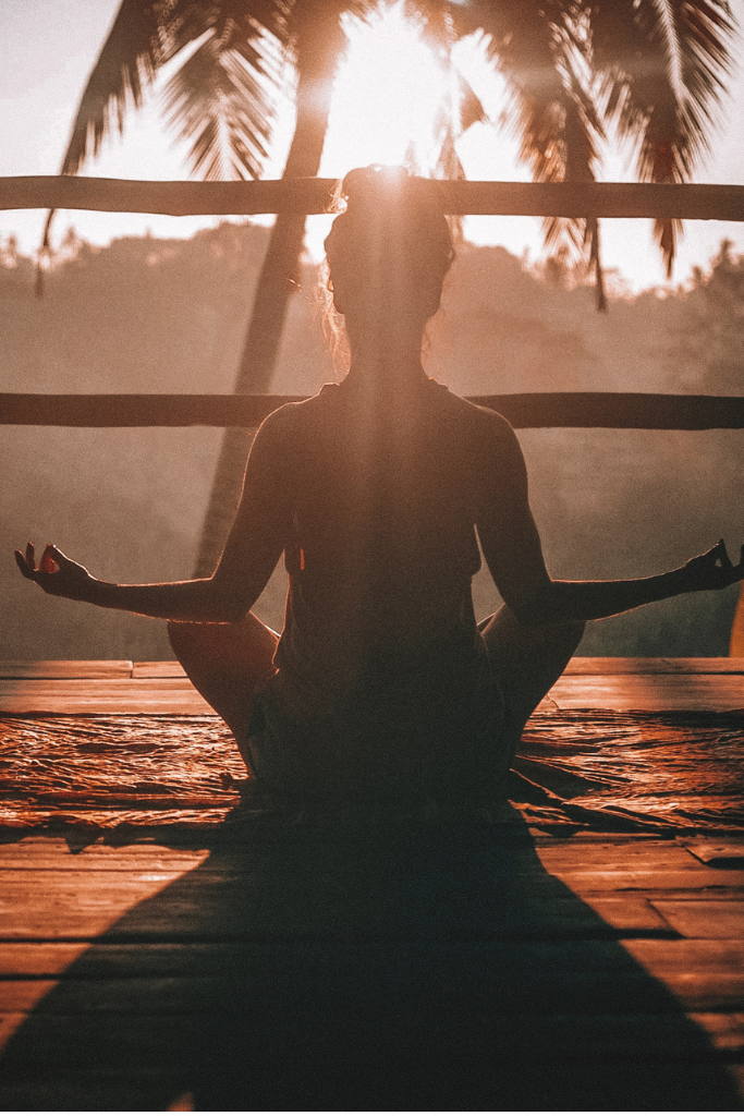 Photo of the back of a woman, on a yoga mat, meditating in front of a palm tree.