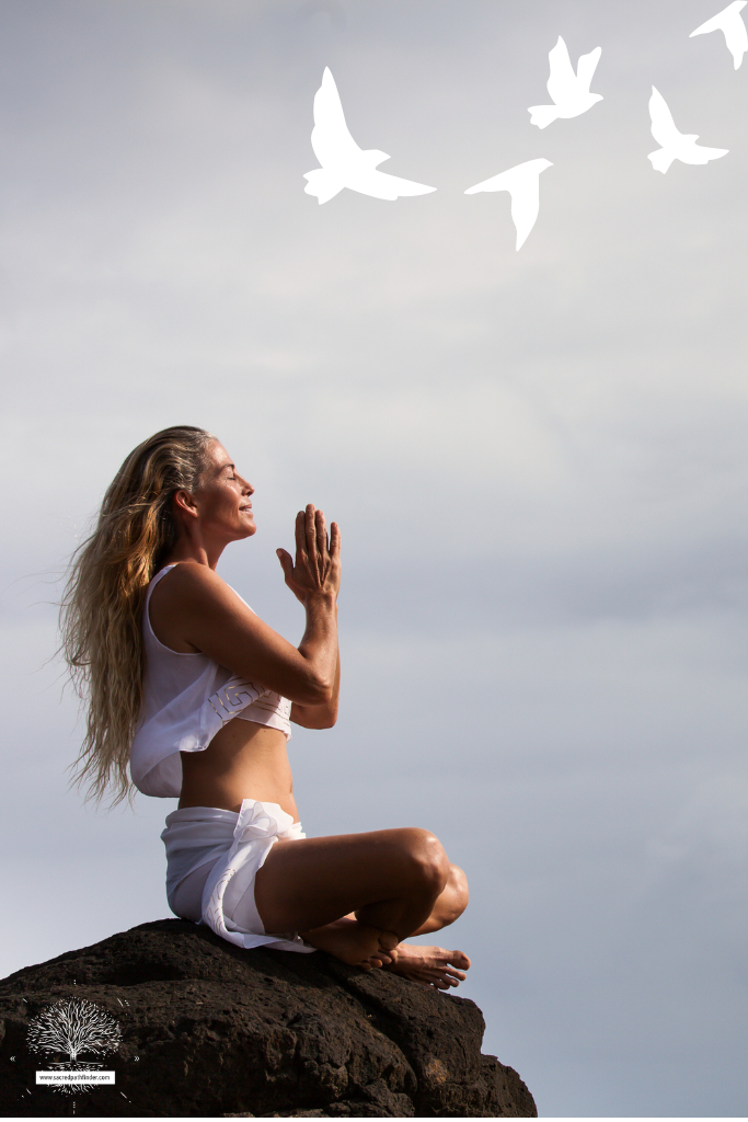 Photo of a woman on a rock cliff, meditating. There are doves flying in the sky. 