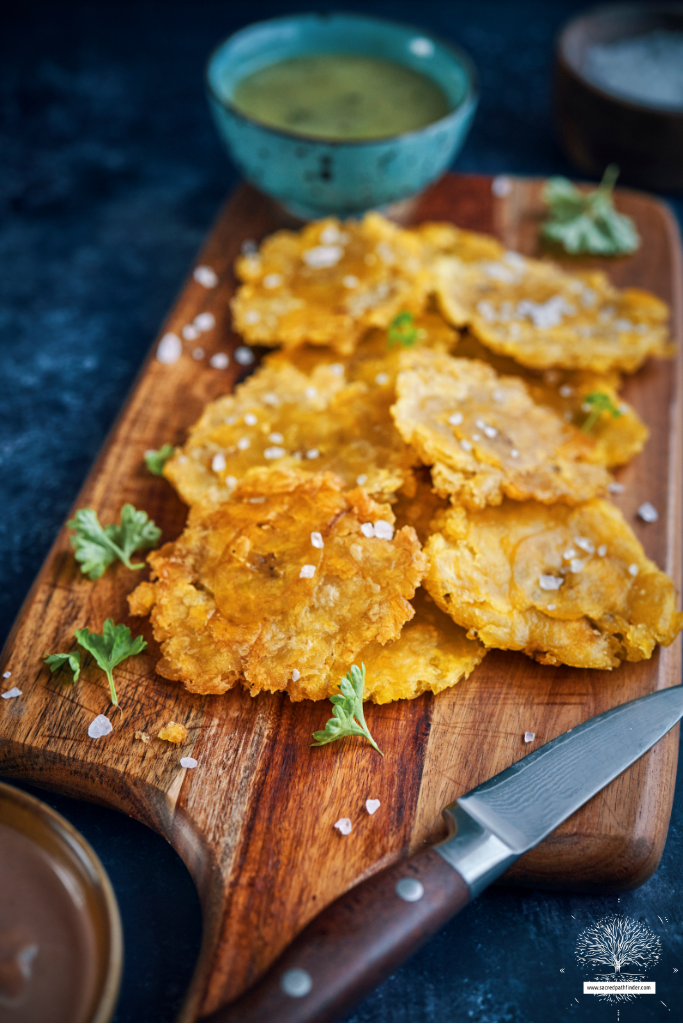 Photo of plantanos on a wooden cutting board. There is a knife in the foreground and tomatillo salsa in the background. 