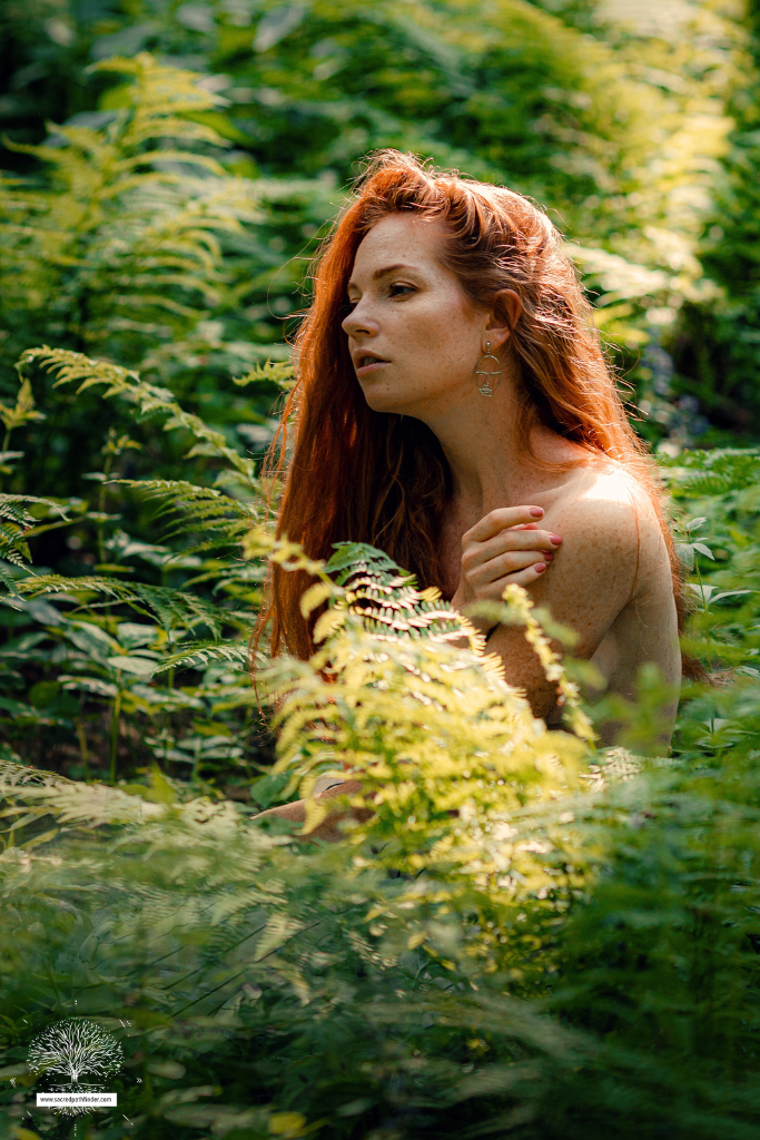 Photo of a woman with long red hair in nature. She is not clothed, but is covered by plants in the picture. 