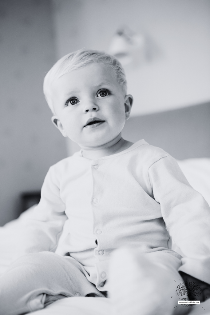 Photo of a baby boy, sitting up on a bed. The photo is black and white. 