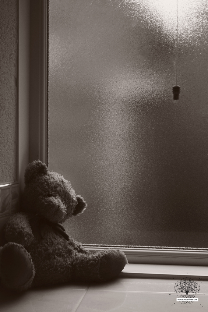 Black and white photo of a teddy bear next to a window.