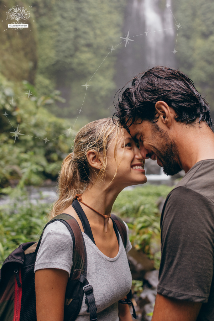 Photo of a couple in front of a waterfall. They are close, touching foreheads. There is an image of the scorpio constellation in the background. 
