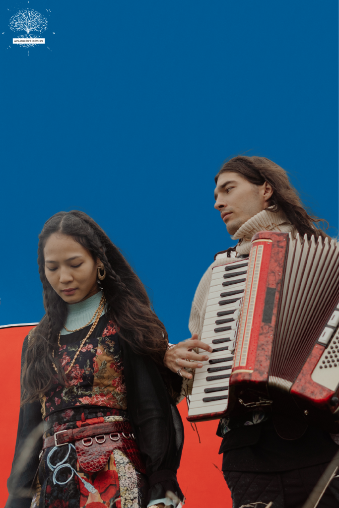 Photo of a couple. The man is playing an accordion for his lady. The background is blue and red. 